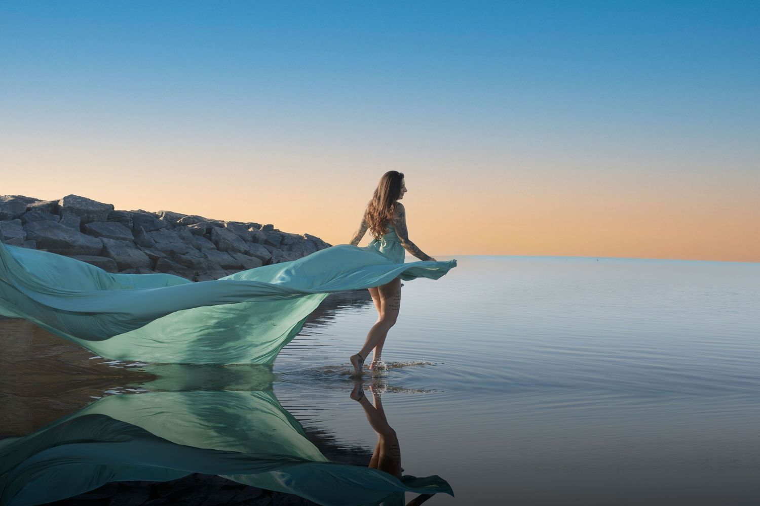 Woman in teal flowing fabric standing in shallow water at sunset near rocky shoreline. Ottawa flying dress photography. Britannia beach