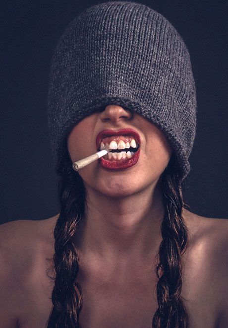 Bold artistic portrait of woman with gray beanie pulled over eyes, red lips, braids, and cigarette.