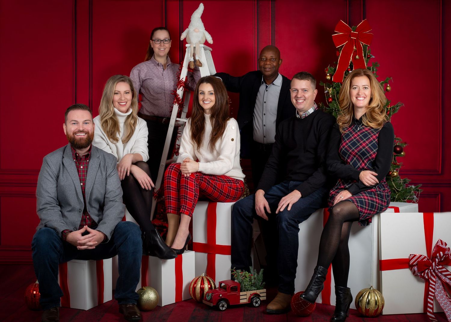 A festive family Christmas photo with seven people posing among wrapped gifts in front of a red backdrop with a decorated tree.