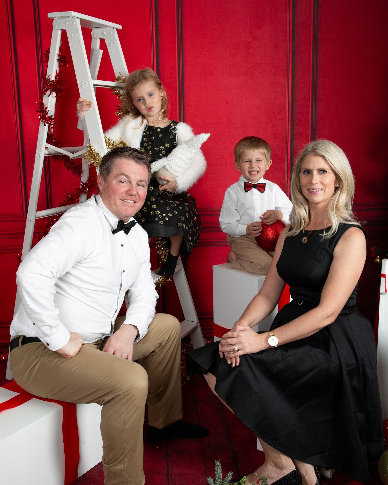 Family of four posing for a festive Christmas portrait against a red backdrop with holiday decorations.