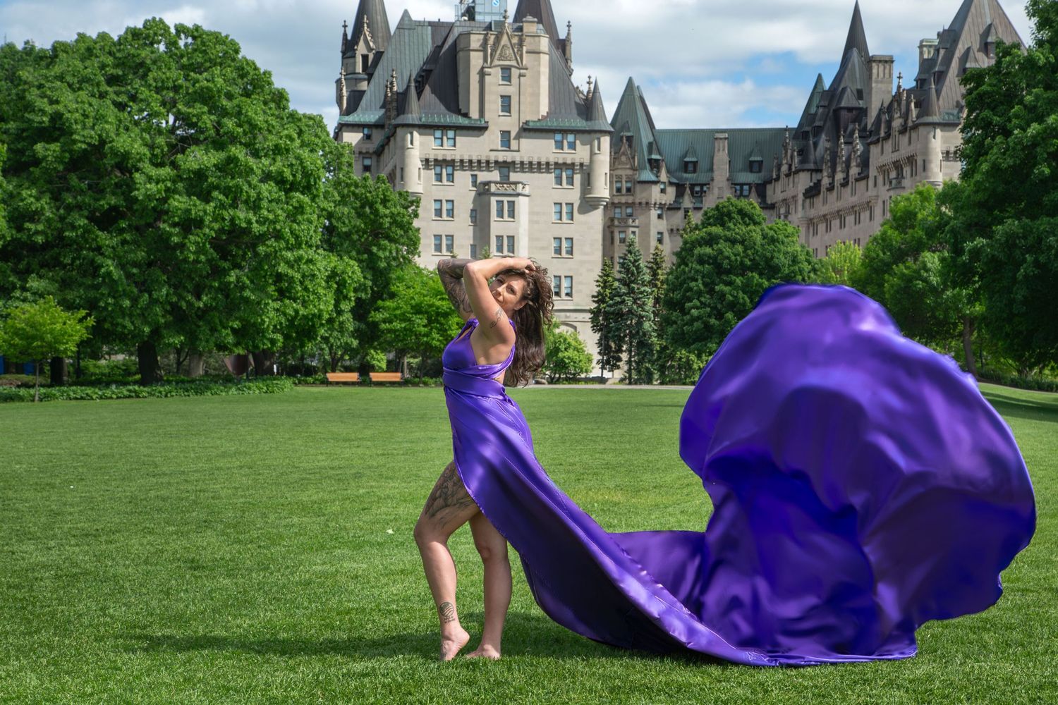 Woman in flowing purple satin gown poses barefoot on lawn with Château Laurier in background. Ottawa flying dress luxury photography