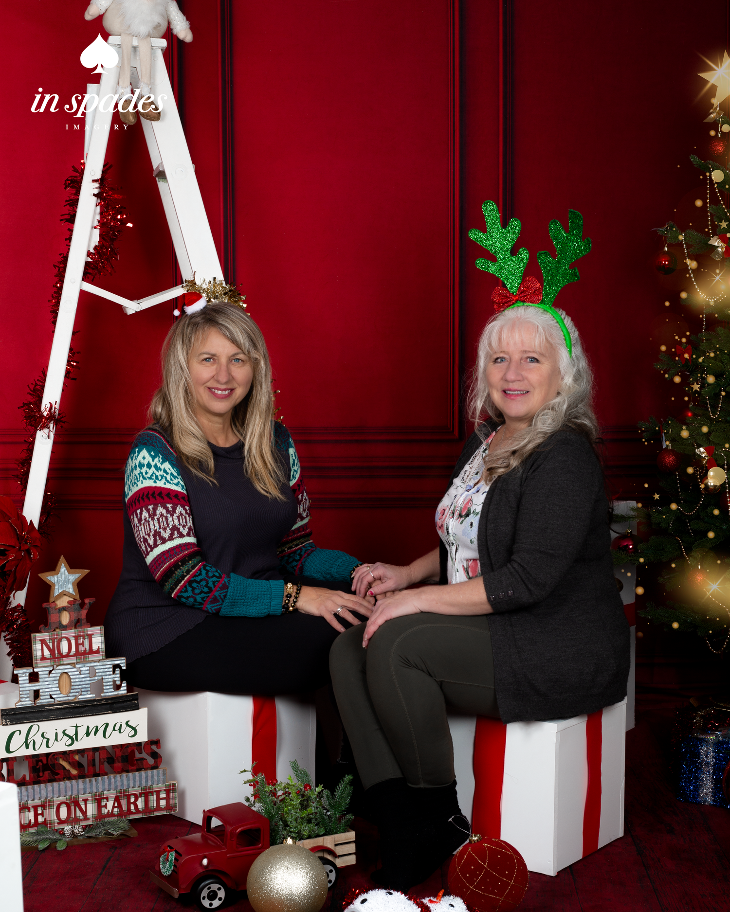 Two women in festive Christmas attire pose smiling in a red holiday studio with a decorated tree and gift boxes.