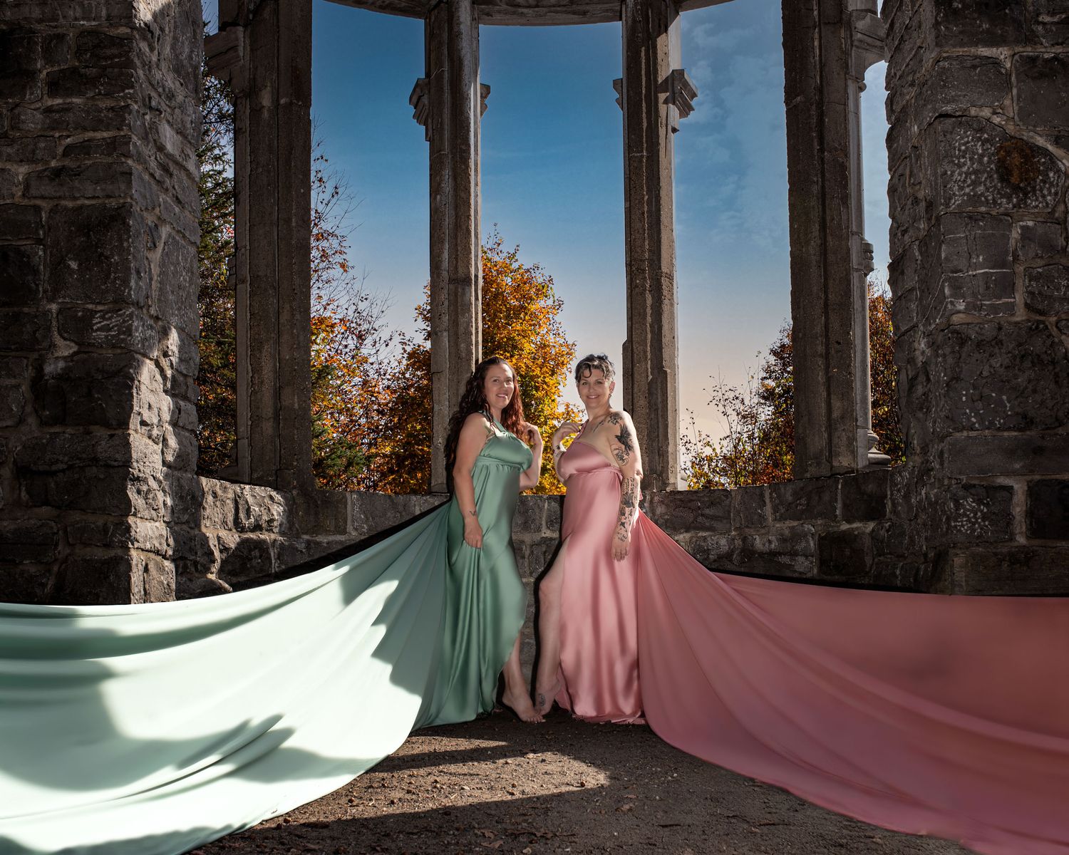 Two women in rose gold sage gowns pose among stone columns with autumn backdrop. Ottawa flying dress photography. Mackenzie King estate