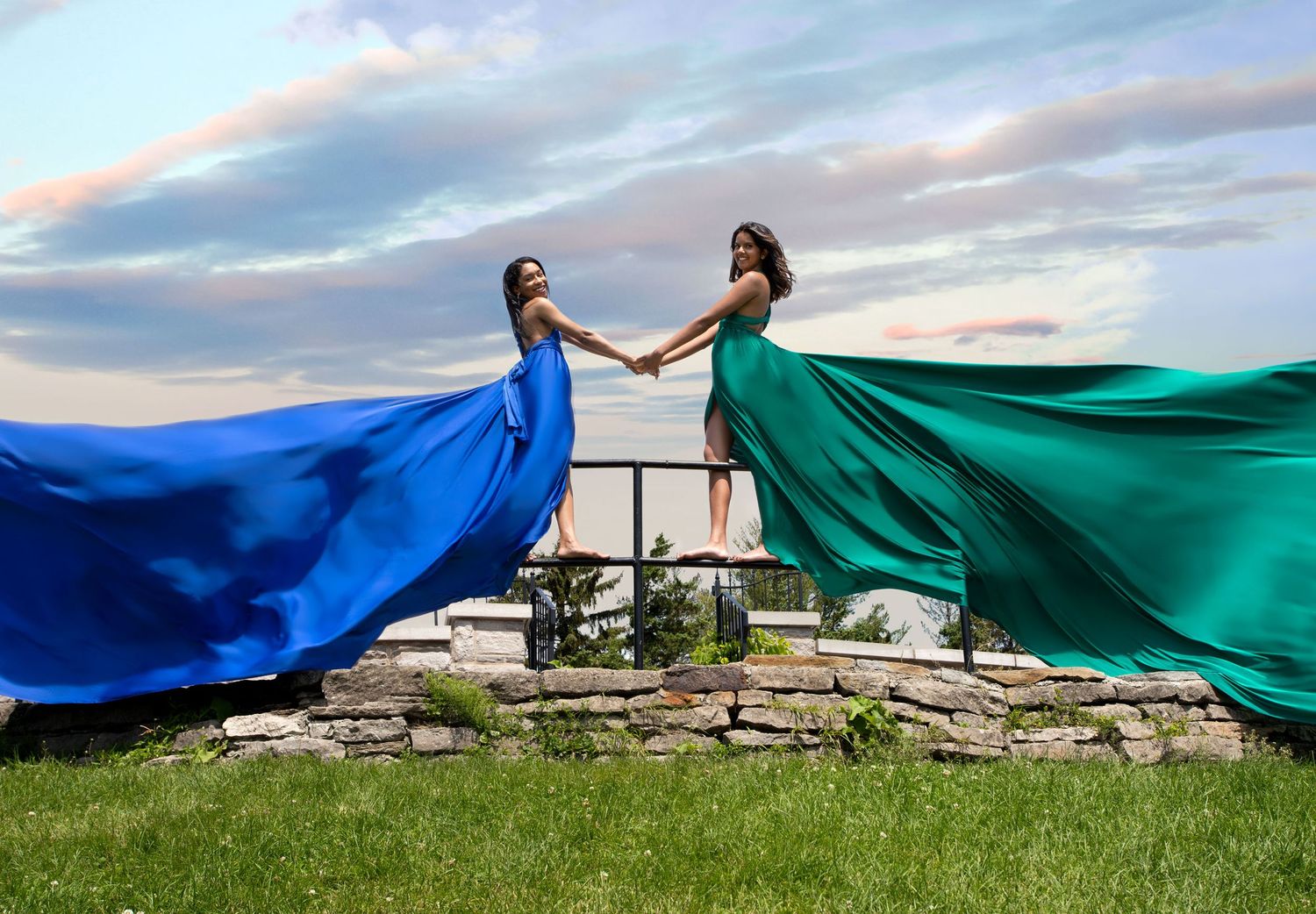 Two women in vibrant blue and green flowing gowns hold hands outdoors at sunset. Ottawa flying dress photography.