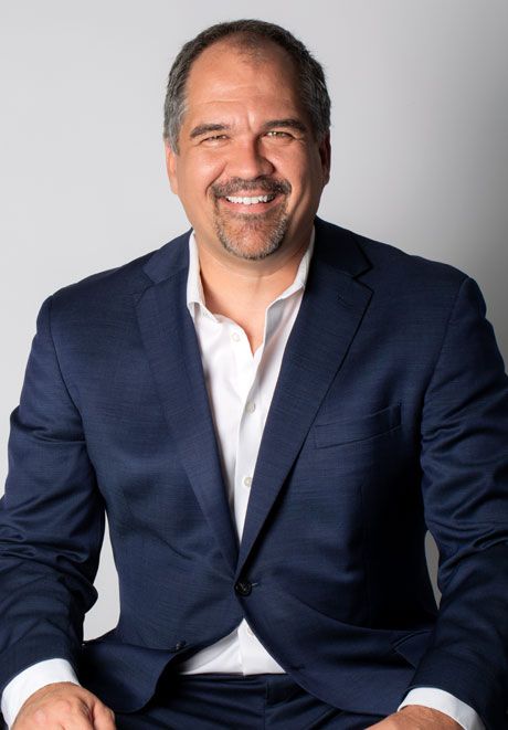 Smiling middle-aged man in navy blazer and white shirt poses for professional headshot against gray background.