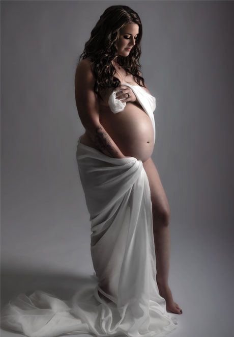Elegant maternity photography of a pregnant woman draped in flowing white fabric against a grey studio background.