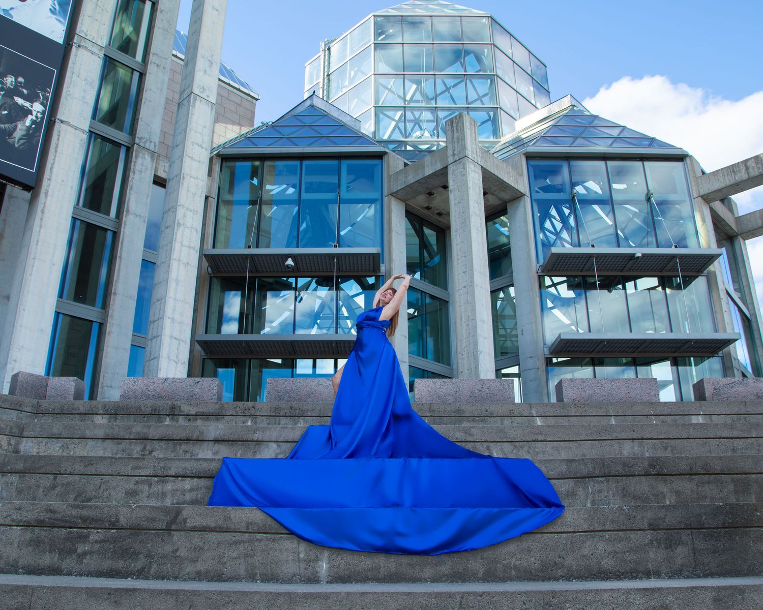 Woman in royal blue flying gown poses on steps before a modern glass building. Ottawa flying dress photography. National Art Gallery