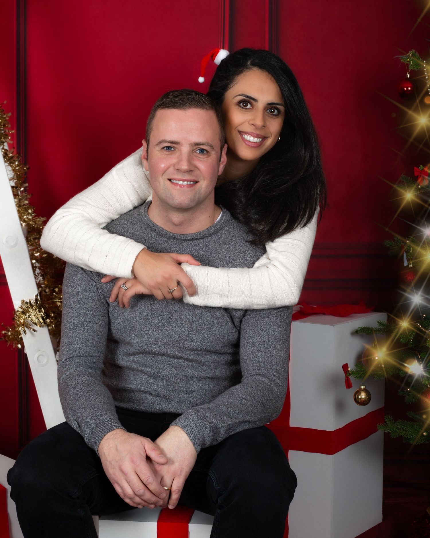 Couple posing for a festive Christmas portrait in front of a red backdrop with gifts and a decorated tree.