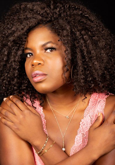 Young Black woman with natural curly hair wears pink lace top and gold jewelry.