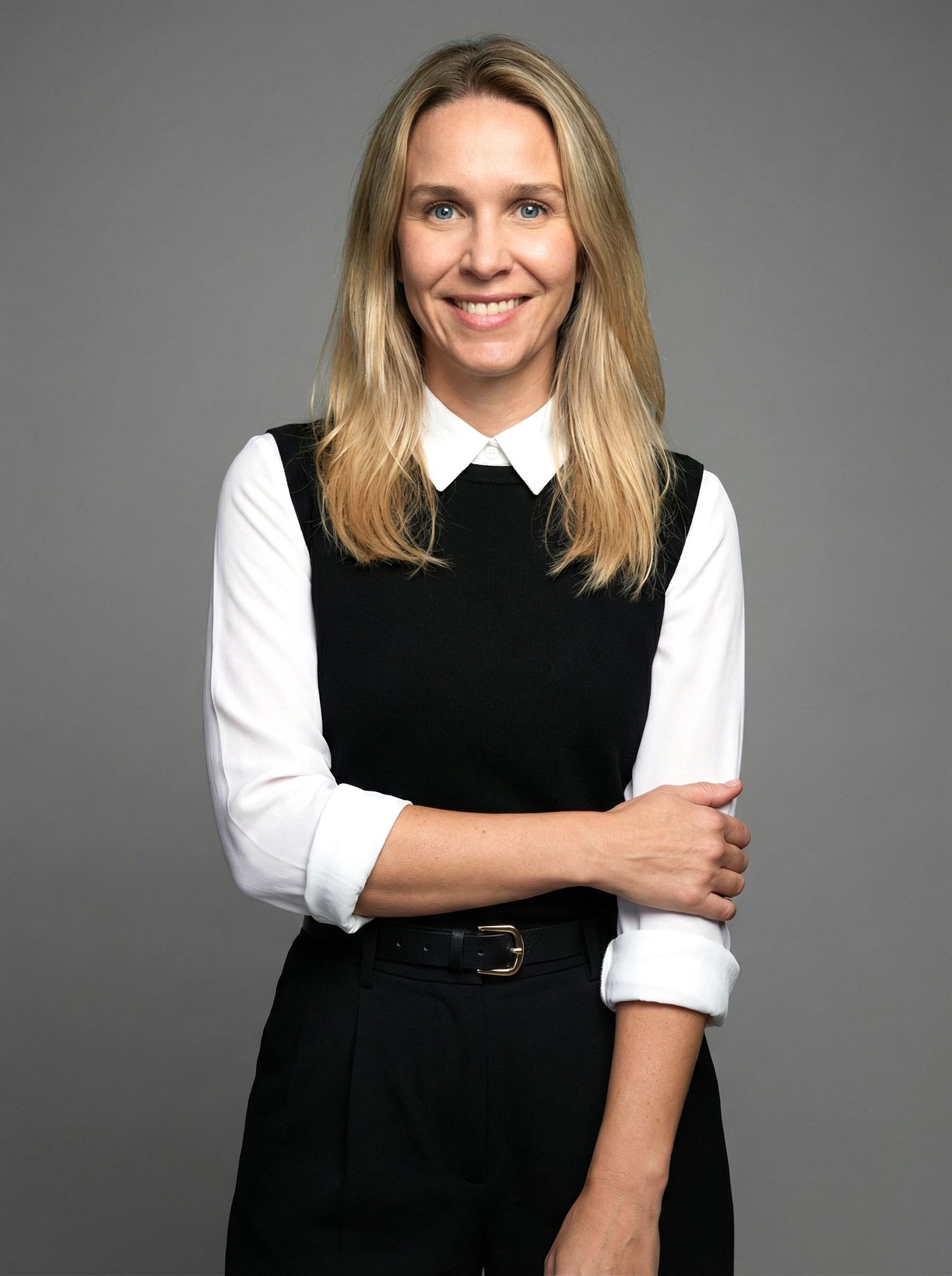 Smiling blonde woman in black vest and white shirt, arms crossed, professional studio portrait.