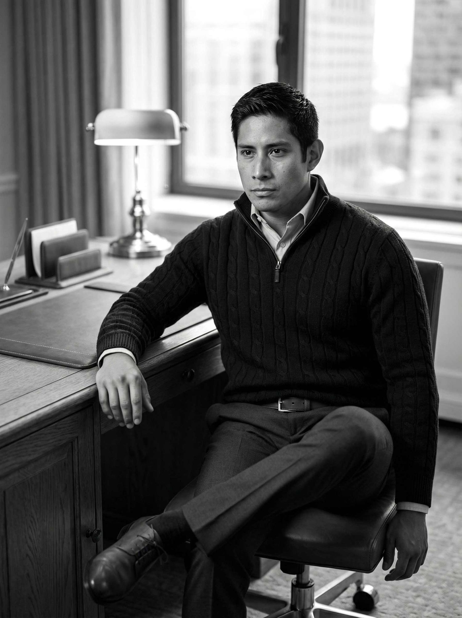 Young man in dark cable-knit sweater sitting at desk, black and white professional portrait.