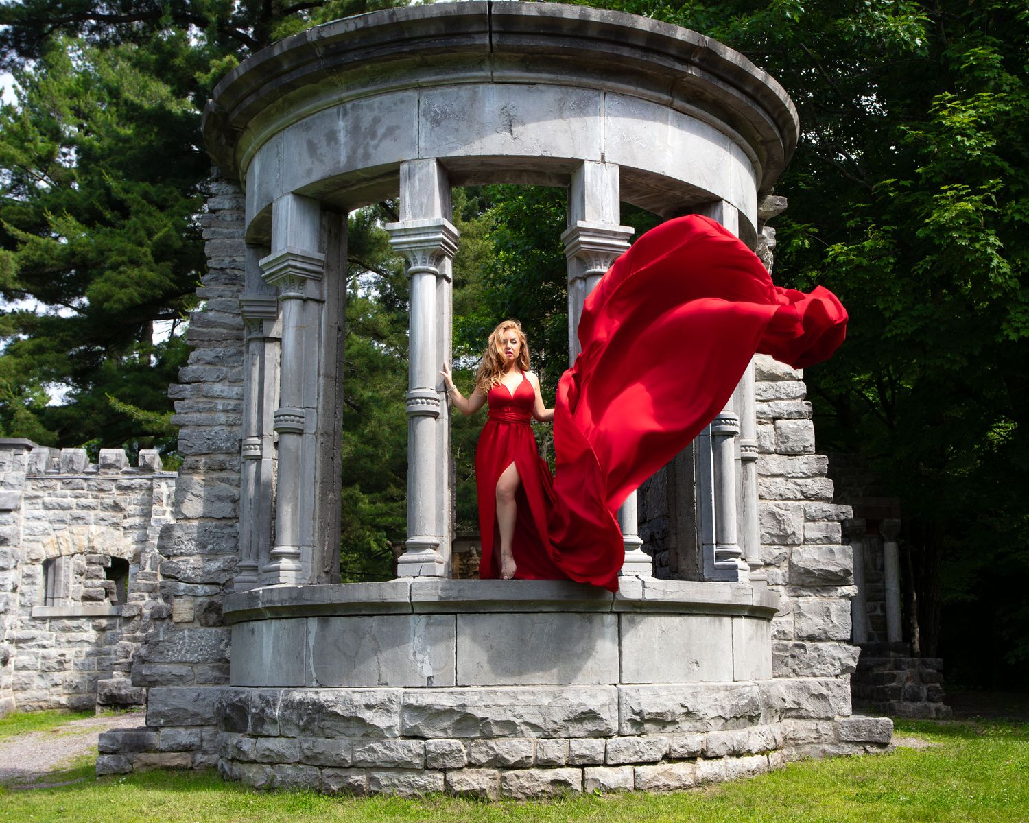 Woman in red flowing gown poses at stone rotunda columns outdoors.  Ottawa flying dress photography. Mackenzie King estate