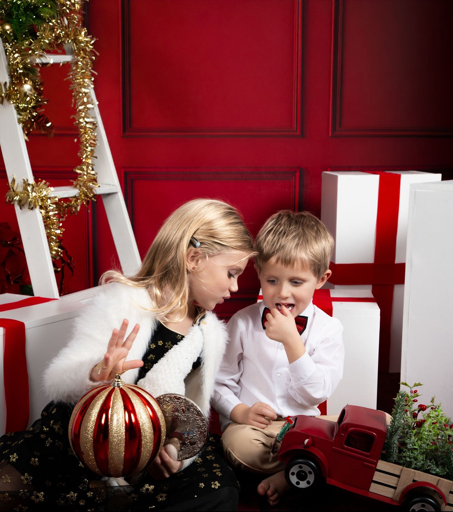 Two children share a joyful moment surrounded by Christmas gifts, ornaments, and festive decorations against a red backdrop.