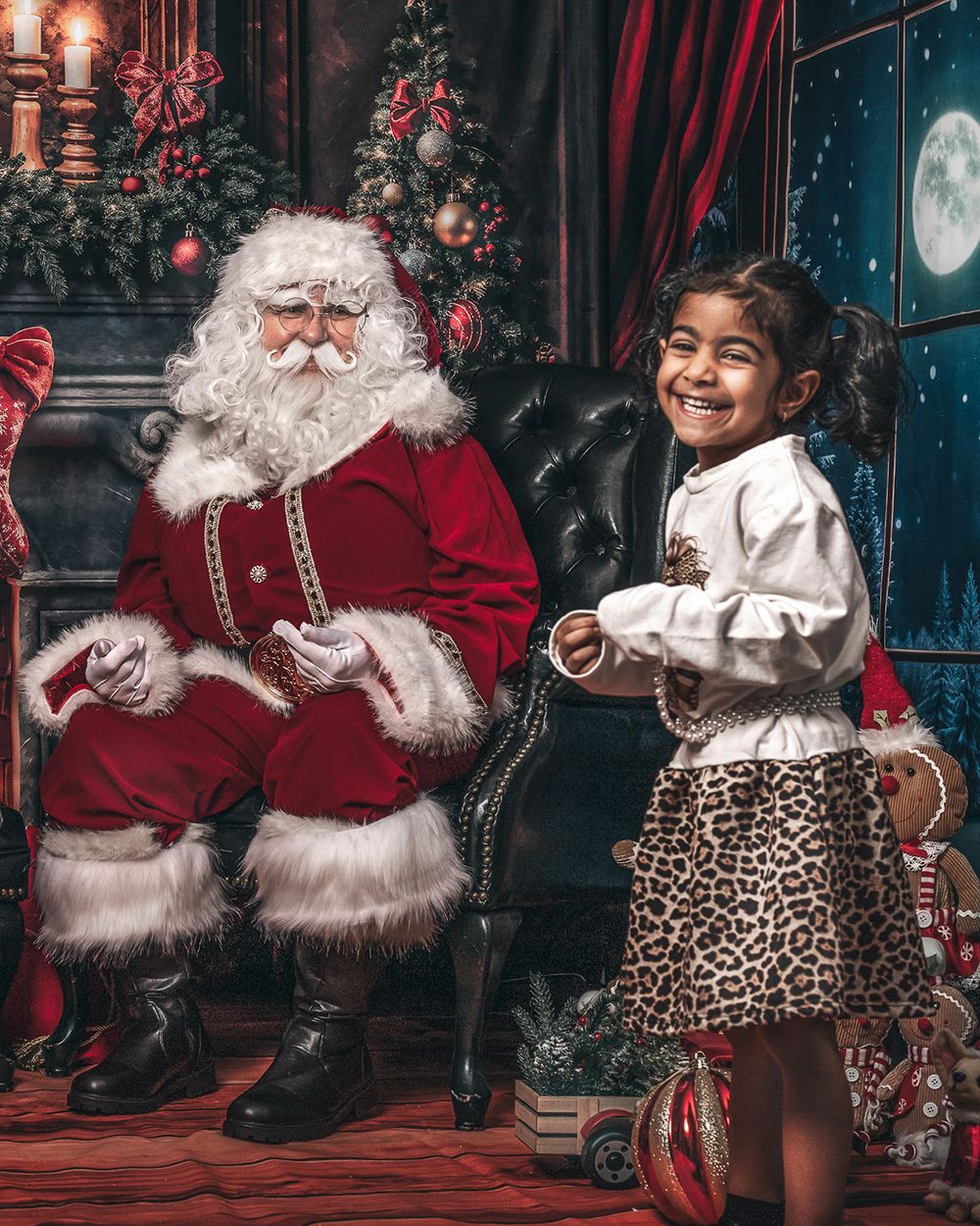 A joyful young girl in a leopard skirt laughs beside Santa Claus in a festive Christmas setting with a decorated tree.