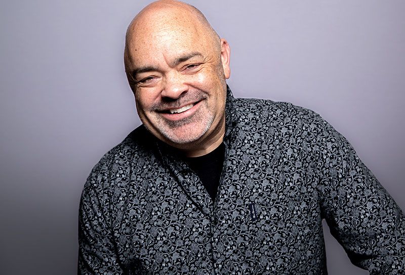Smiling bald man wearing patterned shirt posing against light gray studio background.