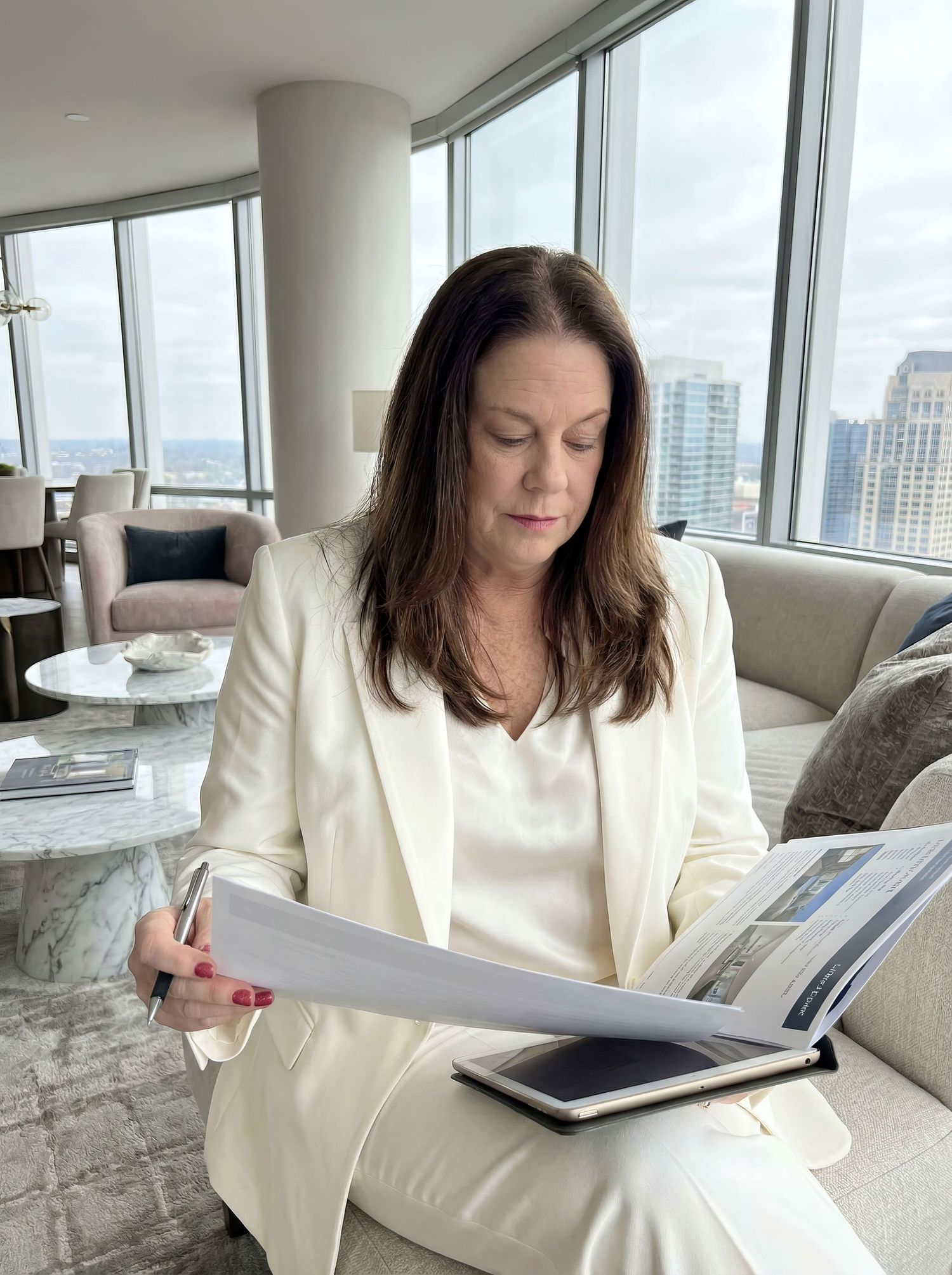 Professional woman in cream blazer reviewing documents in modern high-rise apartment.