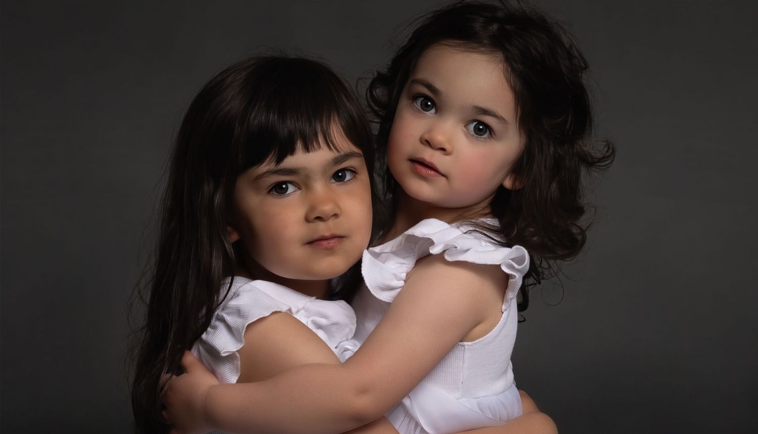 A tender studio portrait of two young dark-haired sisters in white dresses sharing a warm embrace.an Ottawa studio Family portrait.