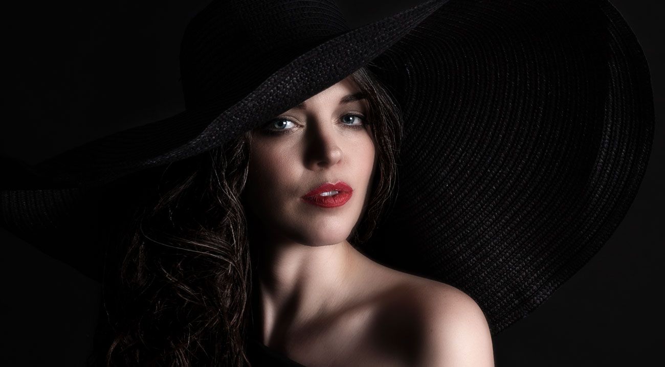 Glamorous woman wearing a wide-brim black hat with red lips on dark background.
