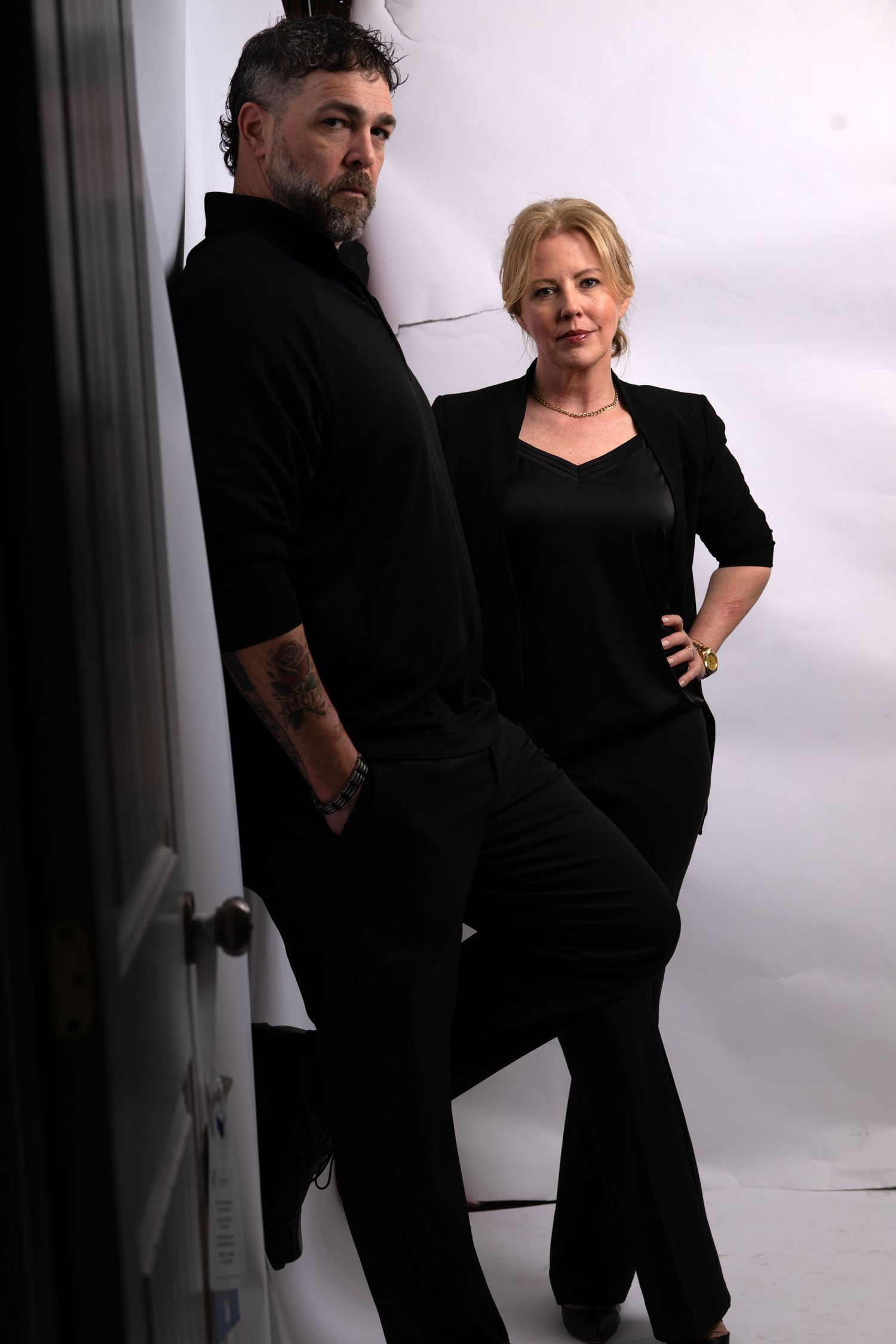 Man and woman in matching black outfits posing against white studio backdrop.