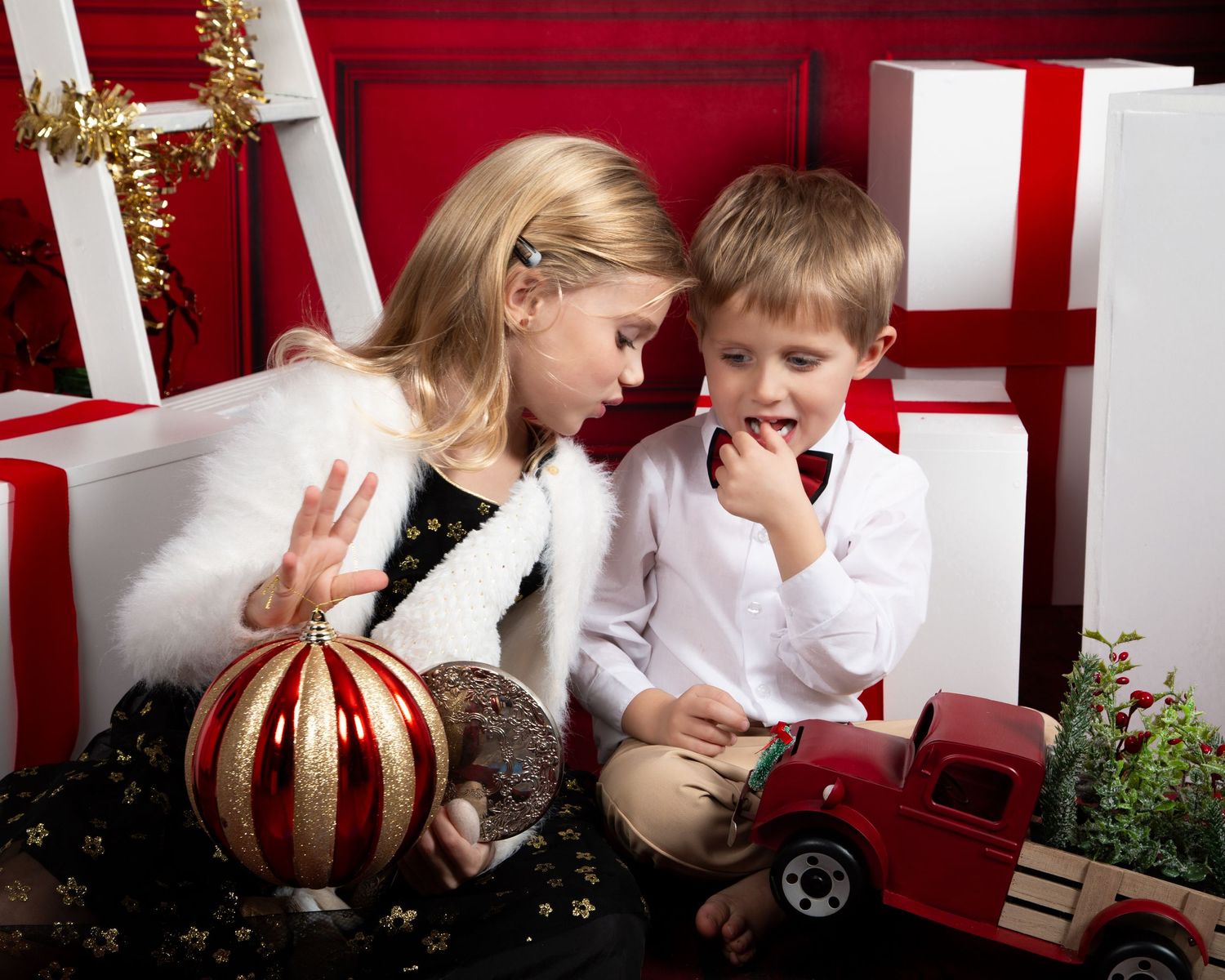 Two children sit together among Christmas gifts and decorations, examining holiday ornaments and a red toy truck.