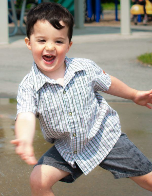 Happy toddler boy in plaid shirt splashing in water puddle at a sunny outdoor playground.