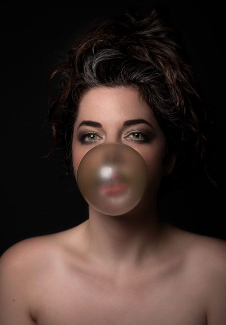 Woman blowing large bubble gum bubble against dark background in artistic portrait.