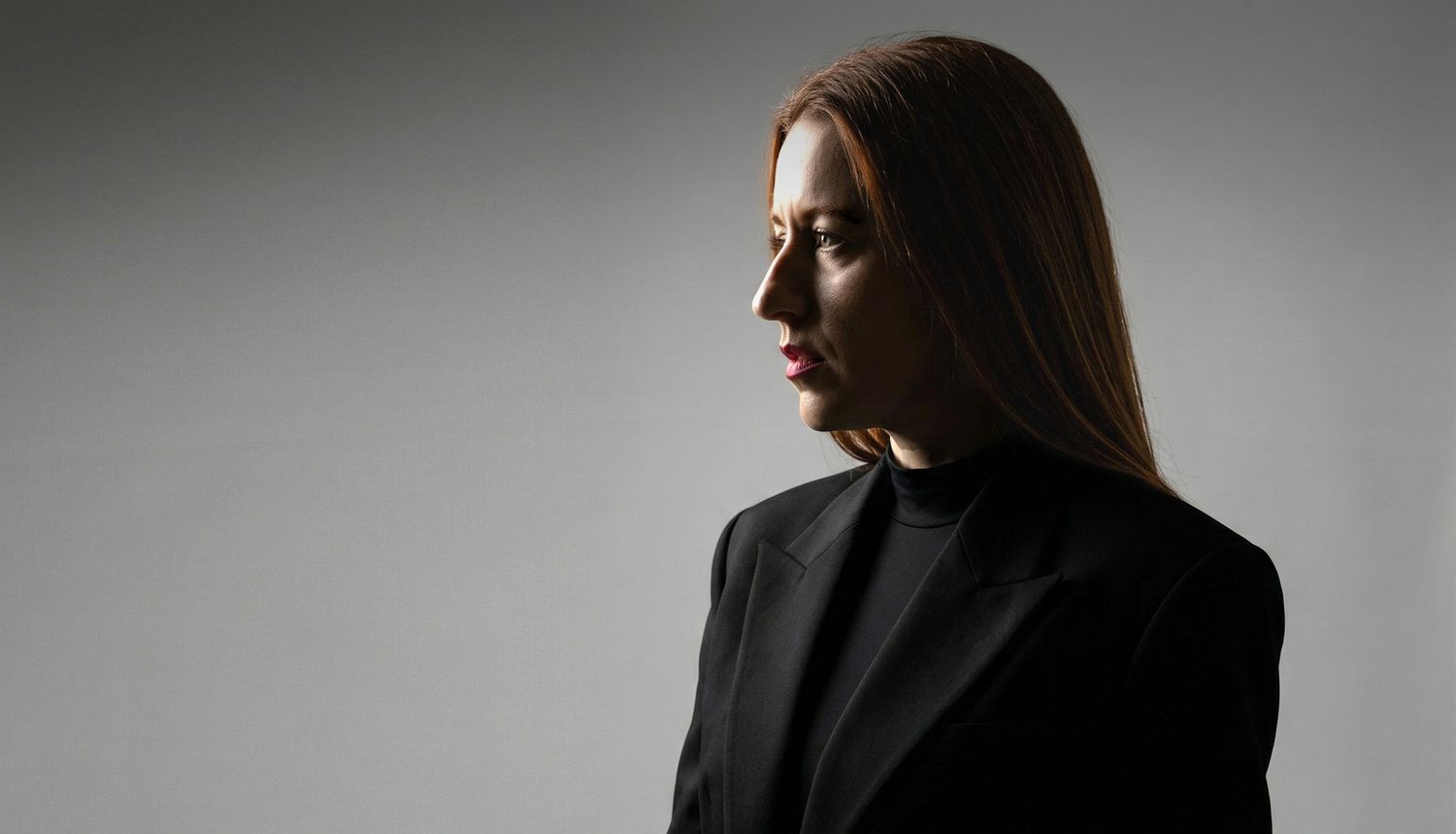 Side profile of a woman with straight brown hair wearing a black blazer against a gray background.