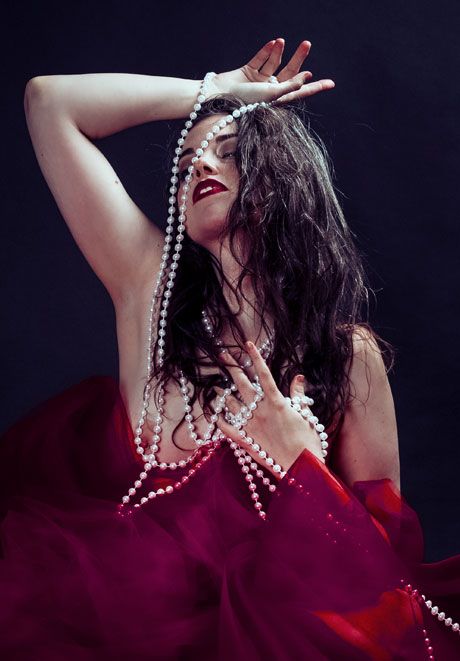 Woman in red dress dances dramatically with pearl necklace against dark background.