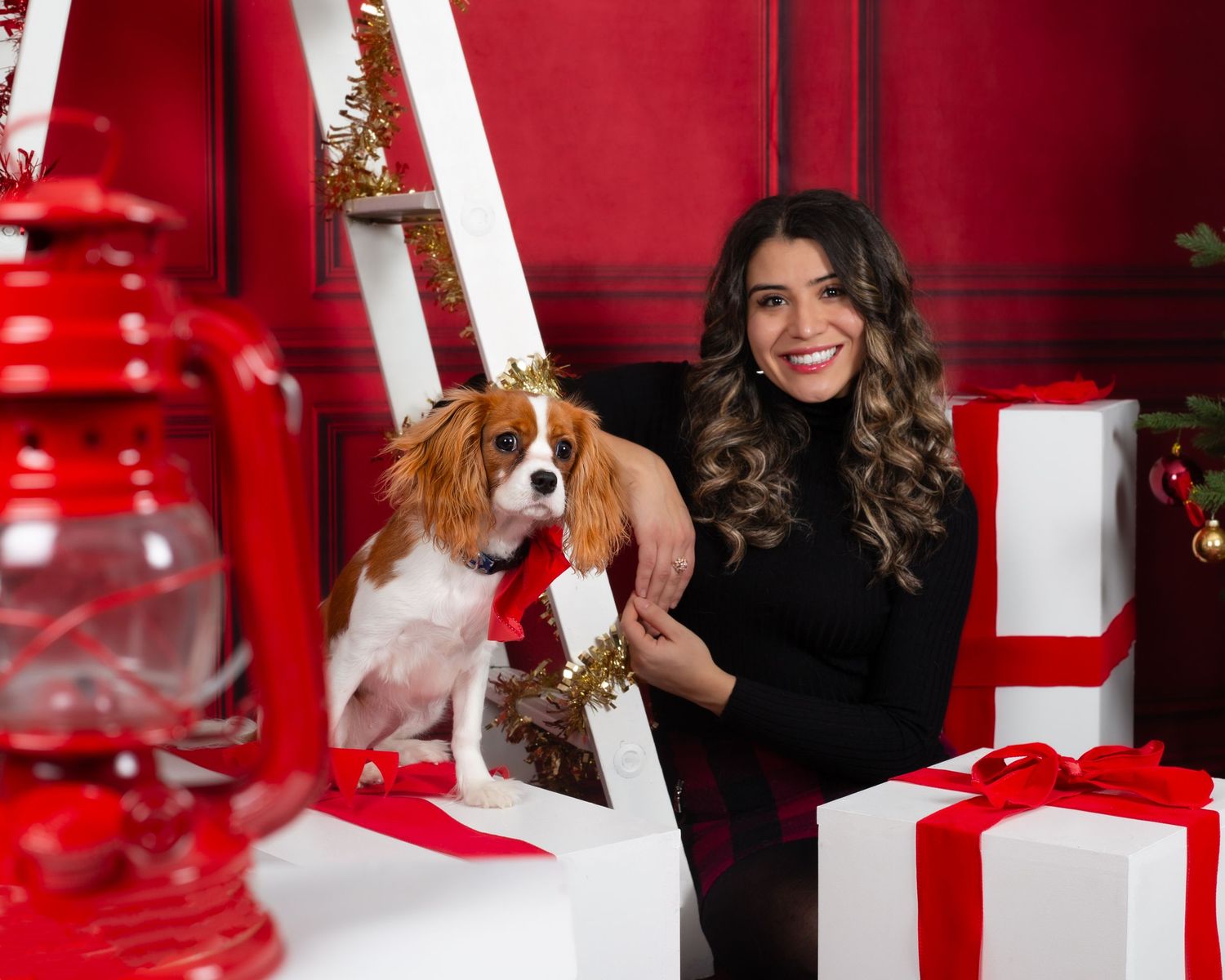 Woman smiling with a Cavalier King Charles Spaniel wearing a crown amid Christmas gifts and red holiday decor.