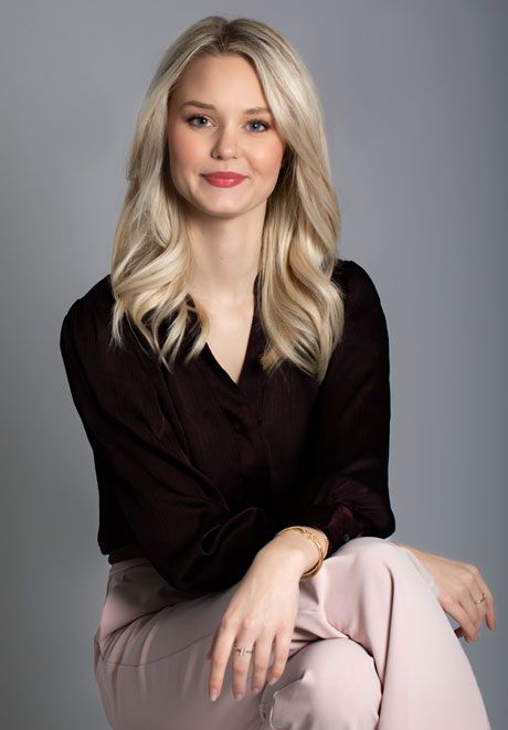 Professional headshot of blonde woman in dark blouse and light pink pants seated against gray background.