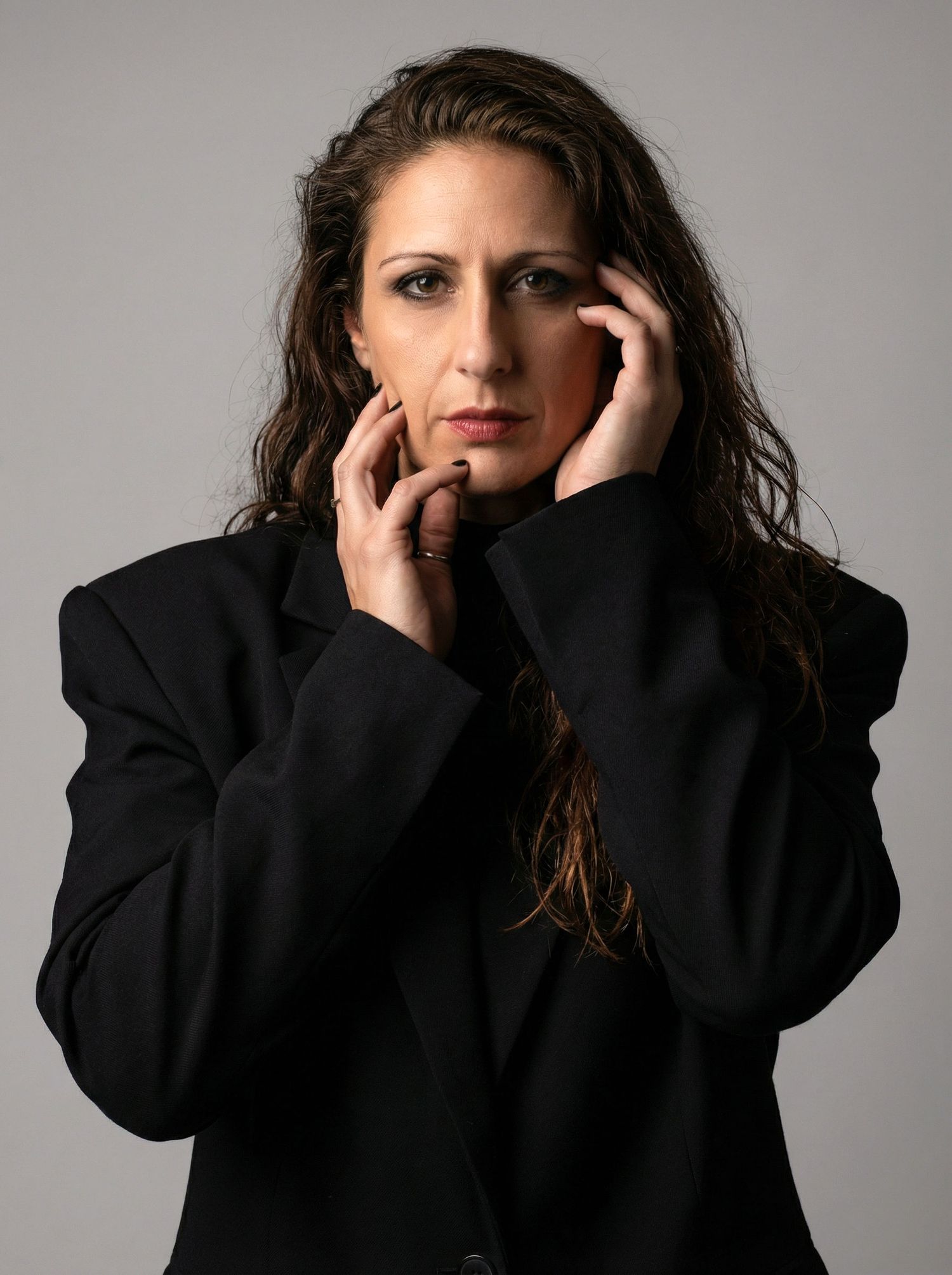 Woman in black blazer holding face with both hands, serious expression, studio portrait.