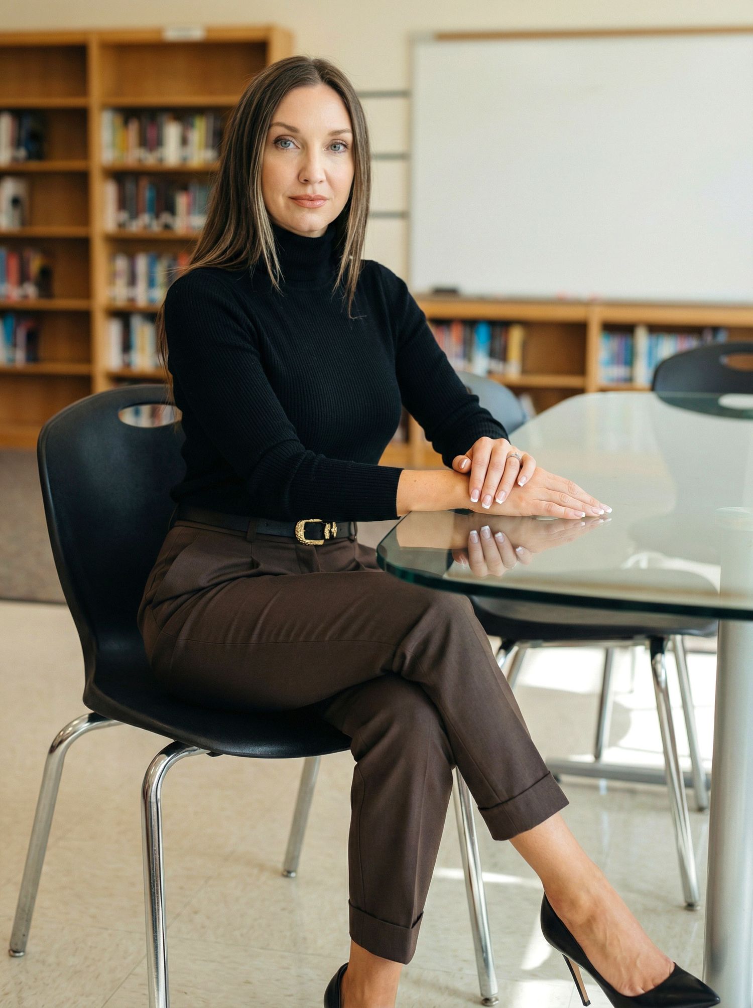 A professional woman in a black turtleneck and brown trousers sits at a glass table in a library setting.