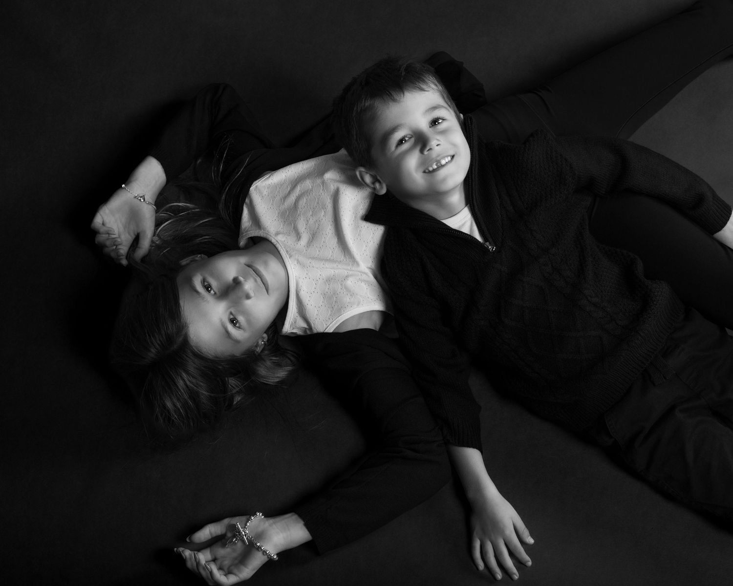 Two young siblings lie on the floor laughing together in a candid black and white studio portrait. an Ottawa studio Family fine art portrait