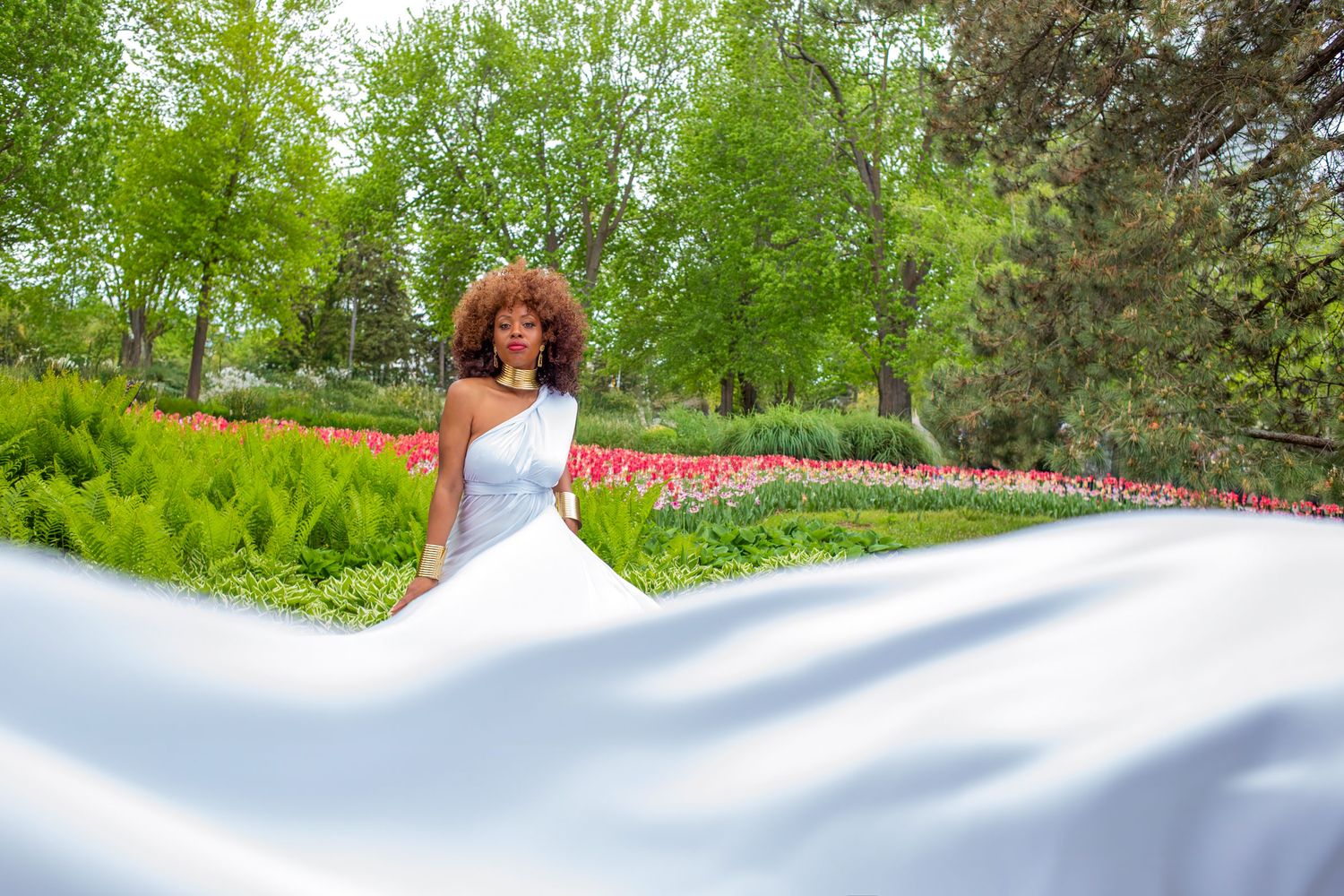 Woman in white flowing gown stands before vibrant tulip festival garden with trees. Ottawa flying dress luxury photography
