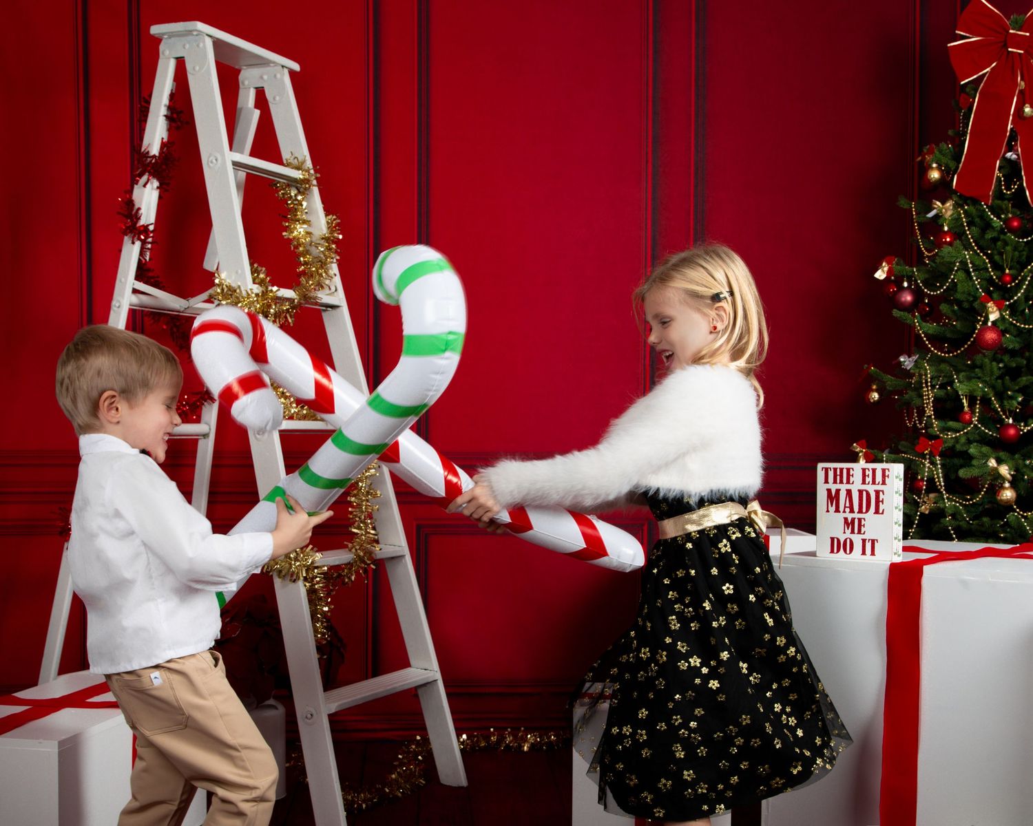 Two children play with oversized candy canes in a festive red Christmas studio with a decorated tree and white ladder.