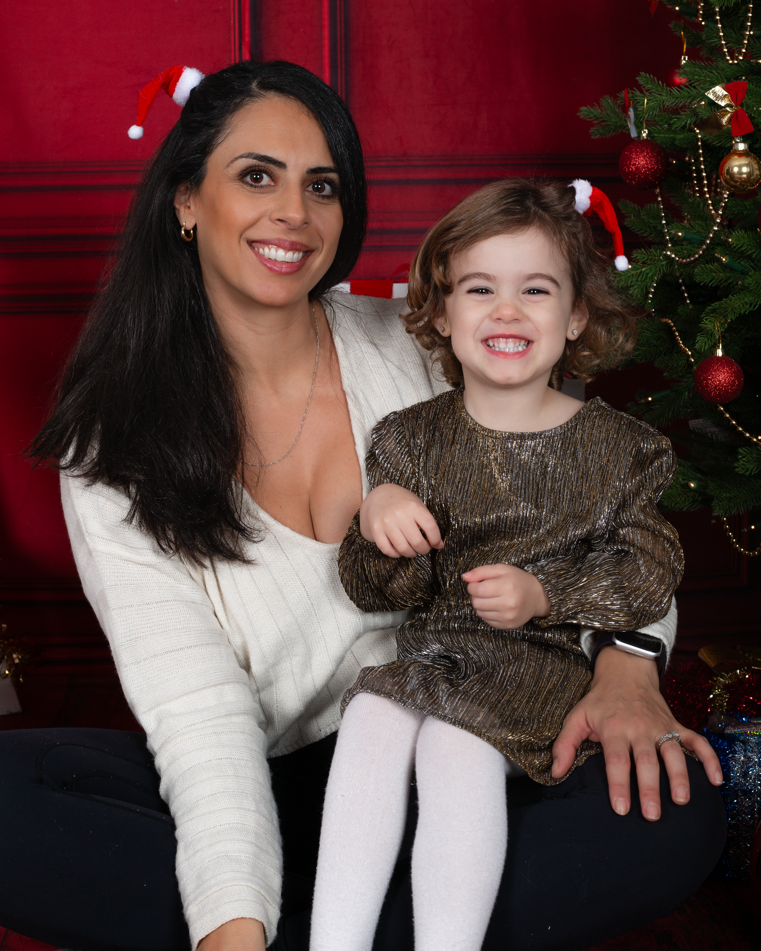Woman and young girl smiling together in front of Christmas tree, both wearing festive outfits for holiday photo.
