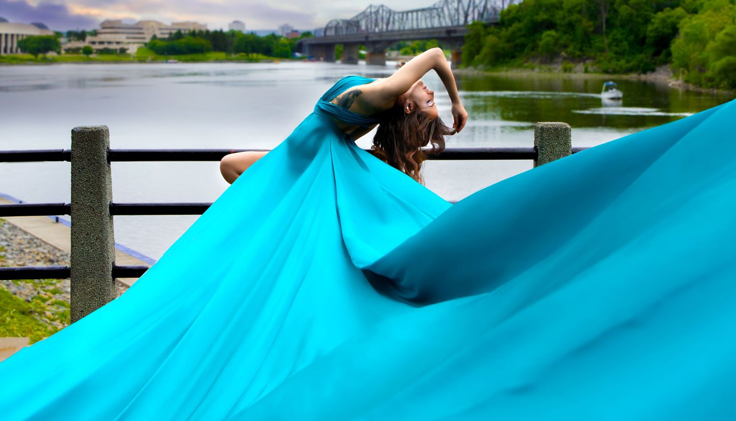 Dancer in flowing turquoise gown posing dramatically by riverside with bridge backdrop.