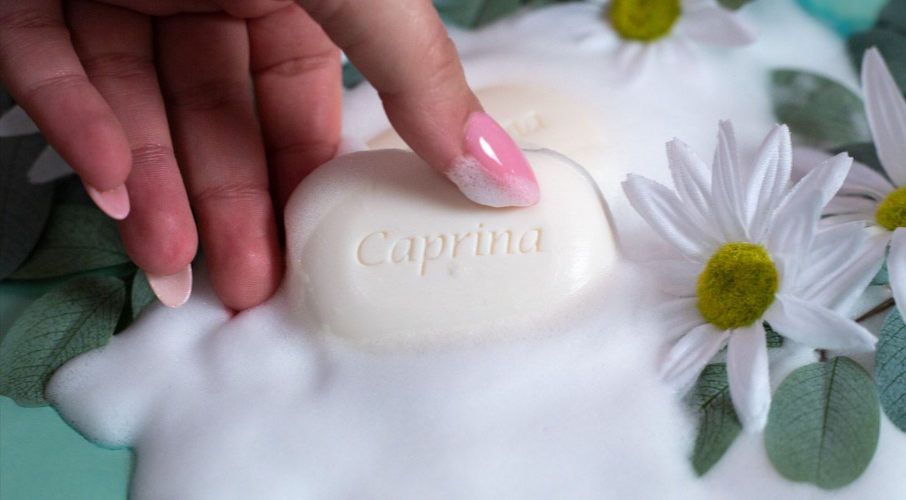 Hand holding Capprina white oval soap bar surrounded by daisy flowers and foam.