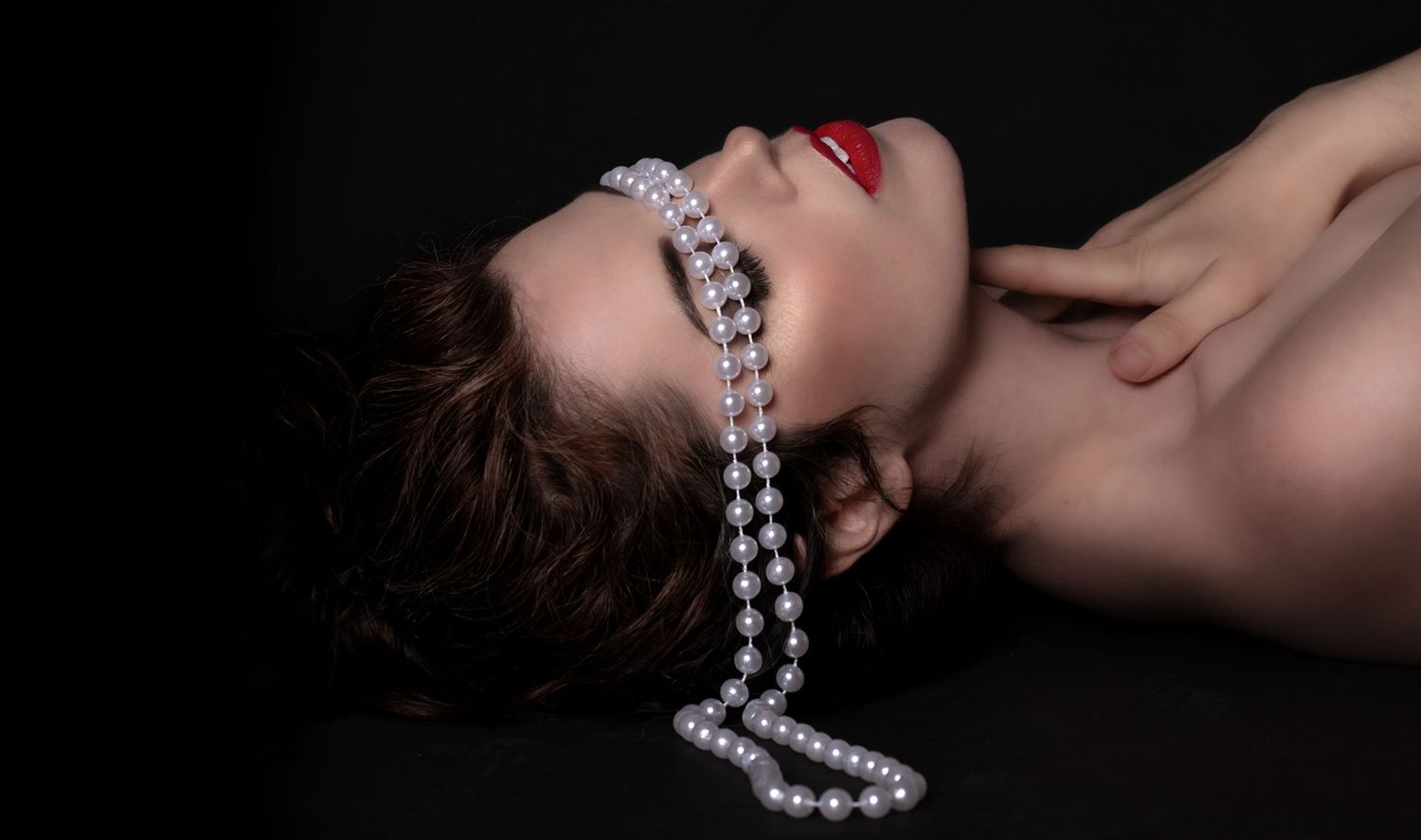 Woman lies back with pearl strand across eyes, wearing red lipstick on dark background.