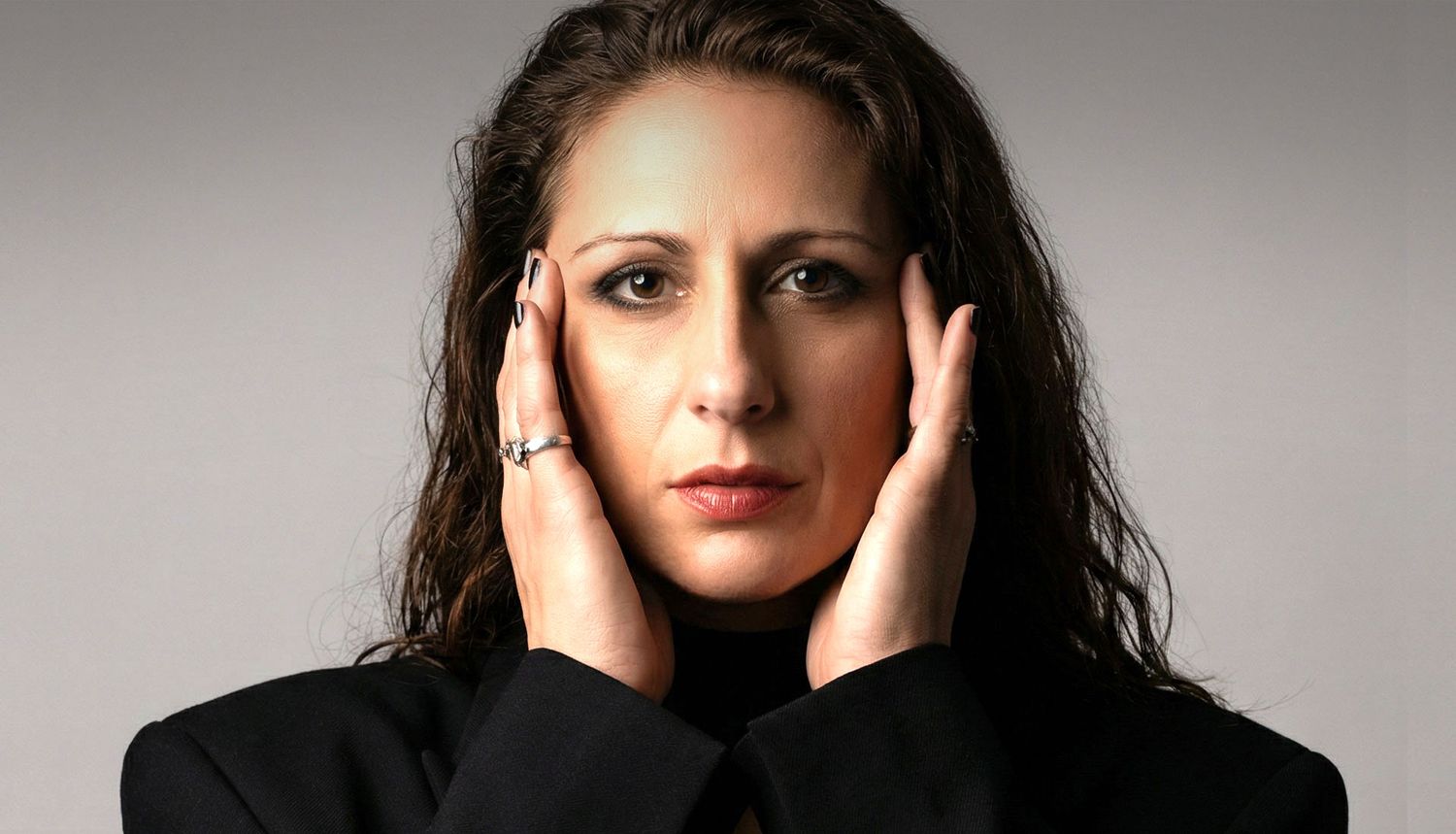 Close-up of woman in black jacket with hands on temples, intense expression, dark background.
