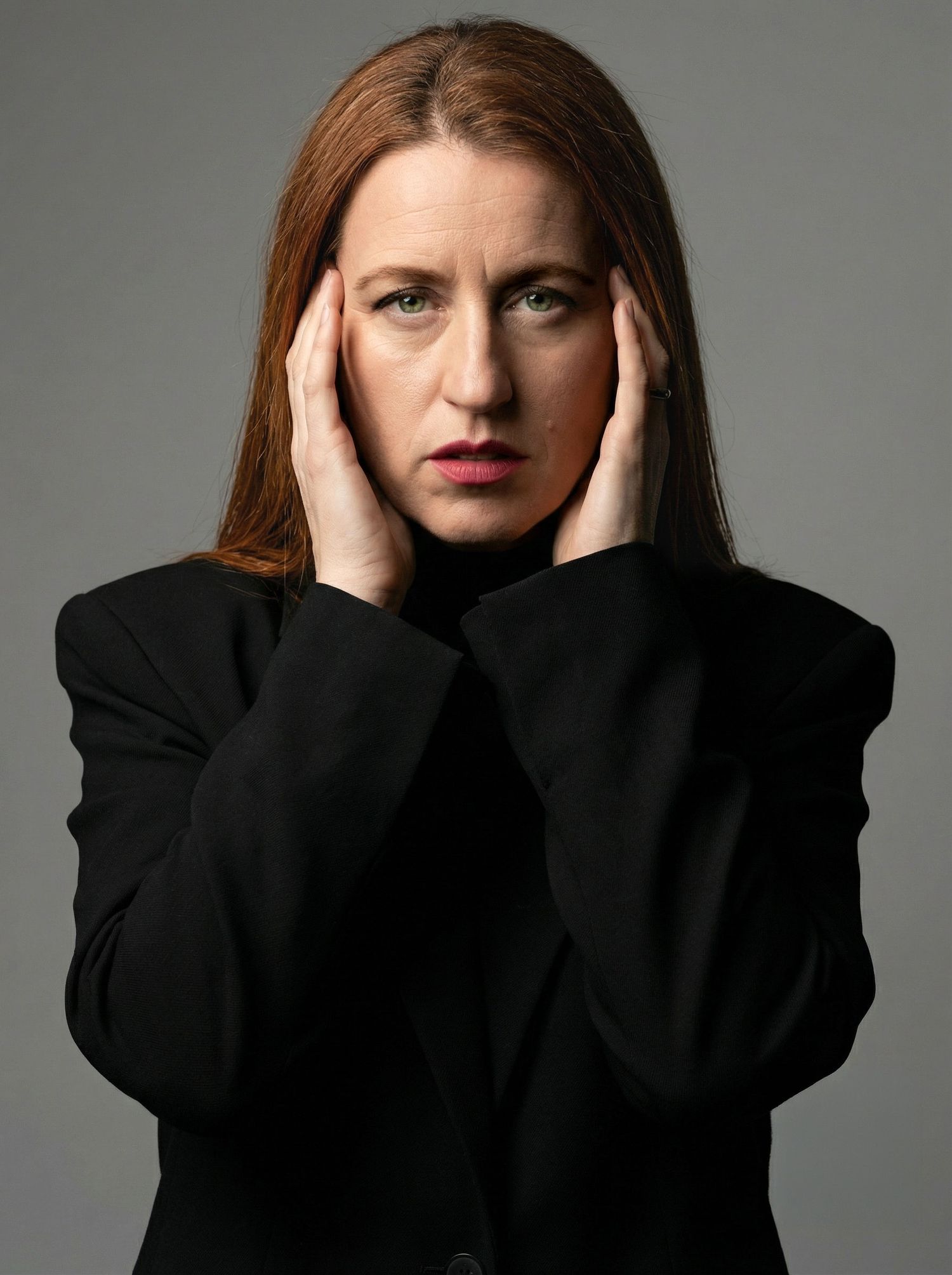 Woman with red hair in black blazer pressing hands to head, stressed look, studio shot.
