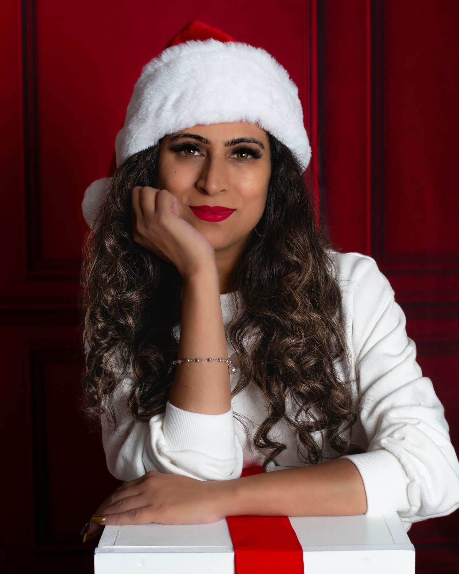 Woman in white sweater and Santa hat poses with gift box against red background.