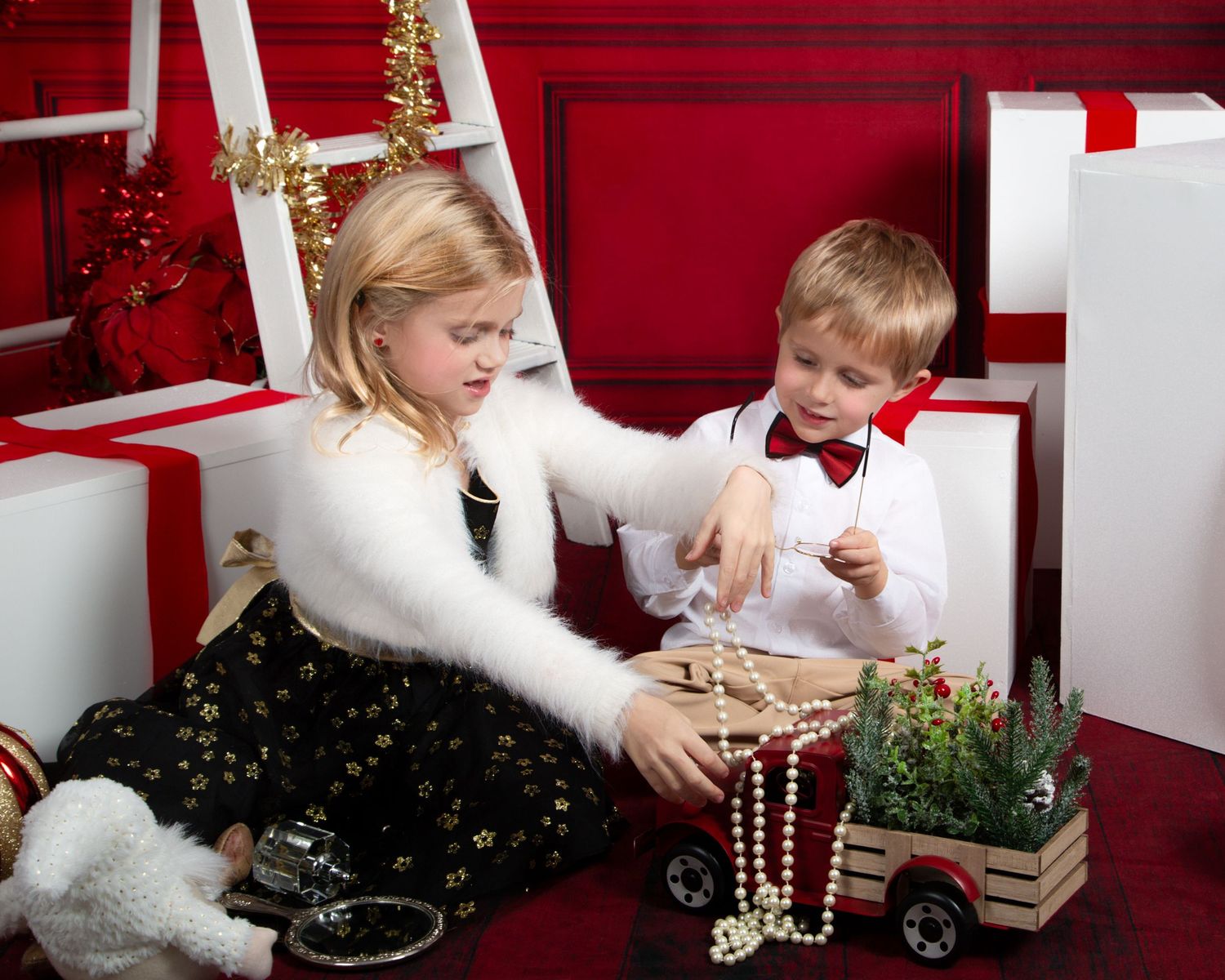 Two young children open Christmas gifts surrounded by white presents and holiday decorations in a festive red studio setting.