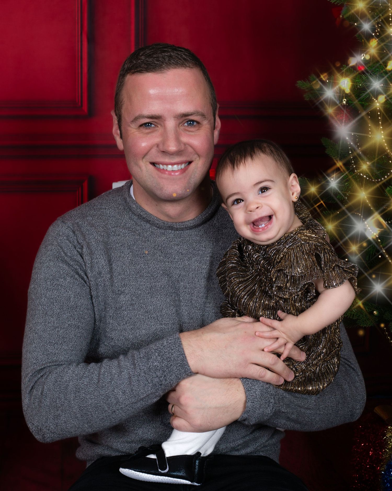Man in gray sweater holding smiling baby in gold dress posing for Christmas portrait near decorated tree.