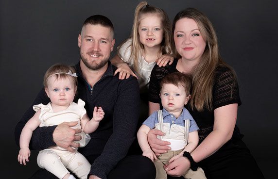 Happy family of five with two parents and three young children posing on dark background.