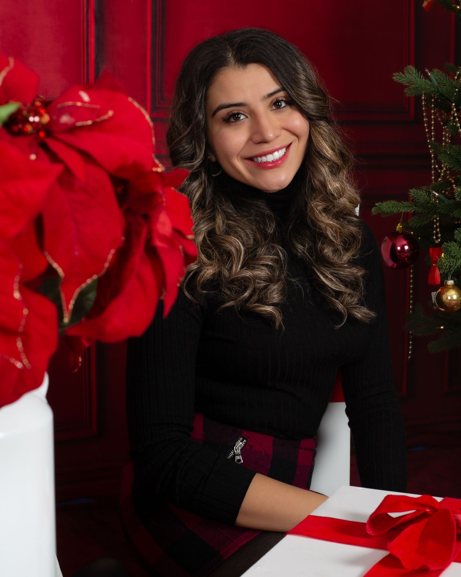 Smiling woman in black turtleneck poses with Christmas gifts, poinsettias, and holiday decorations against a red background.
