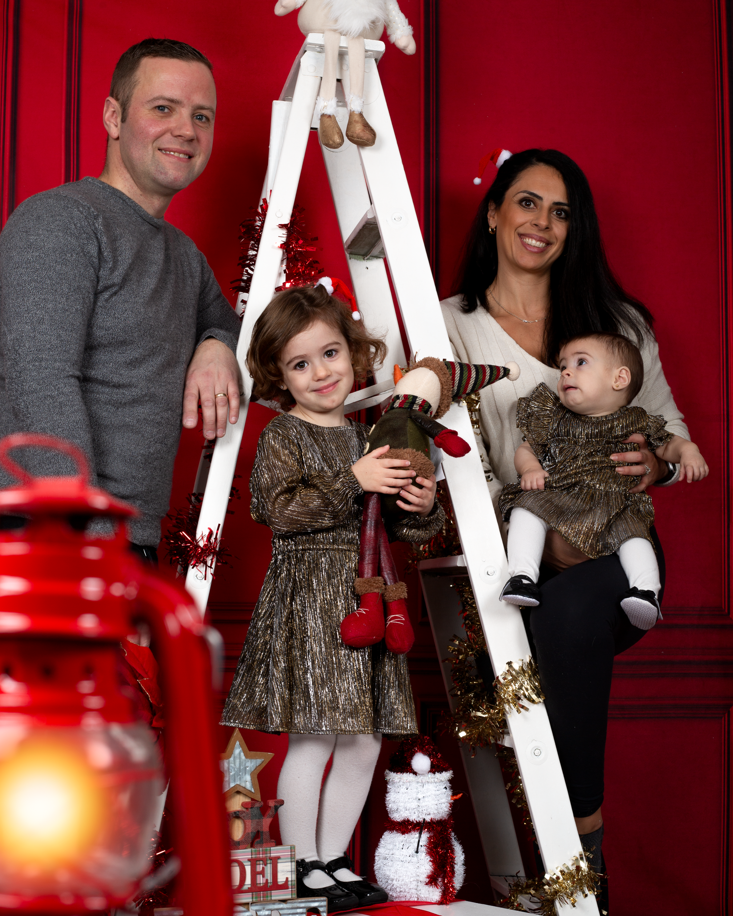 Family of four posing for a festive Christmas photo with a white ladder, holiday decorations, and red background.