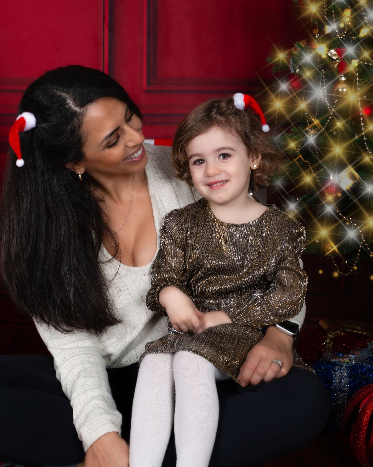 Woman and young girl in Christmas hats sitting together by a decorated holiday tree in festive outfits.