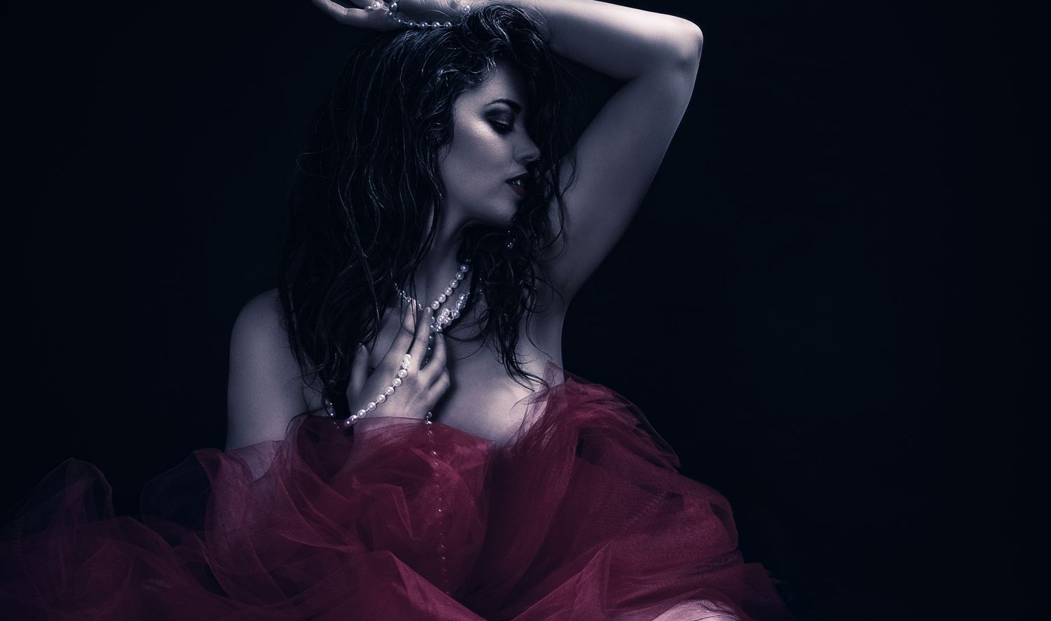 Dramatic portrait of woman in red tulle dress with pearl necklace posed against dark background.