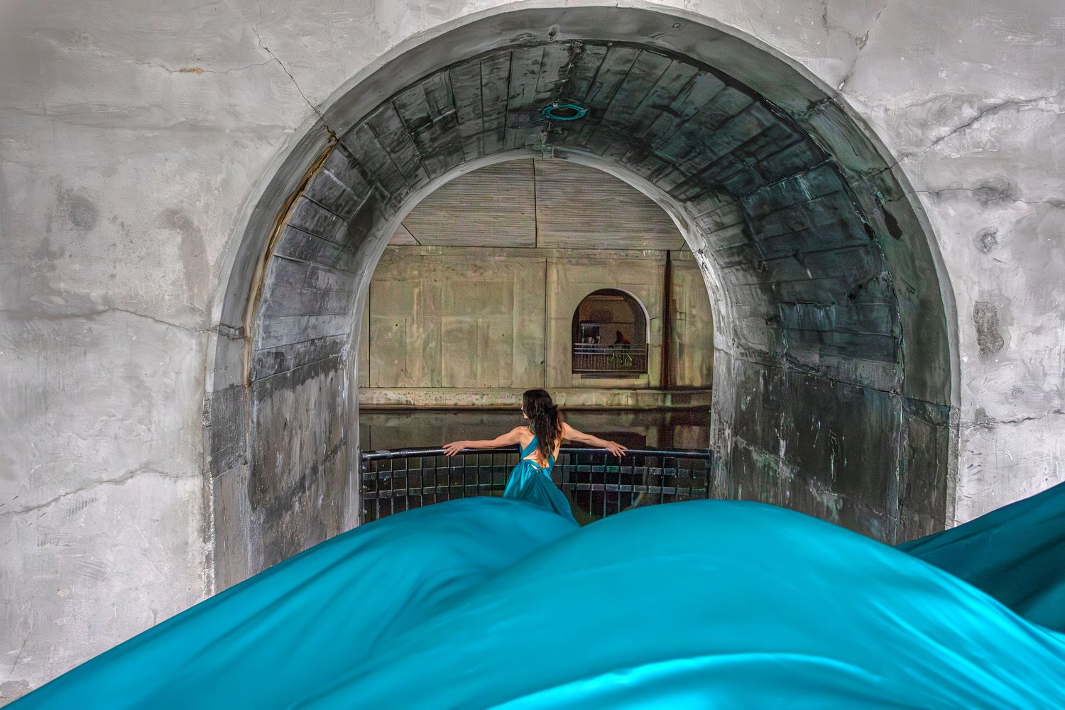 Woman in teal flying dress stands beneath historic stone archway tunnel. Ottawa flying dress luxury photography. Over pass